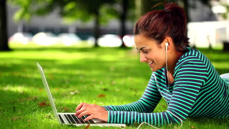 Pretty-student-lying-on-grass-using-laptop-listening-to-music