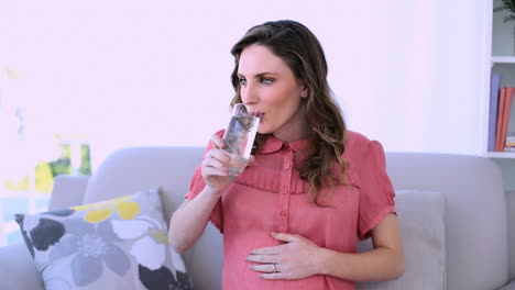 Pretty-pregnant-model-sitting-on-her-couch-drinking-glass-of-water