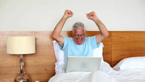 Senior-man-sitting-in-bed-using-laptop-and-cheering