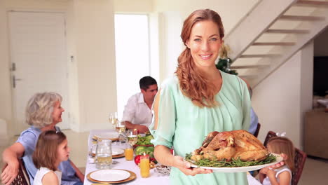 Happy-mother-showing-roast-turkey-to-camera-with-family-behind-her