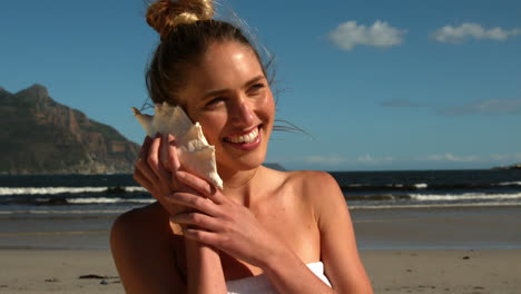 Lächelnde-Blondine-Hört-Muscheln-Am-Strand