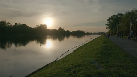 Sunset-reflecting-on-a-calm-lake-with-people-walking-along-the-lakeside-path-at-Jarun-Lake,-Zagreb-Croatia