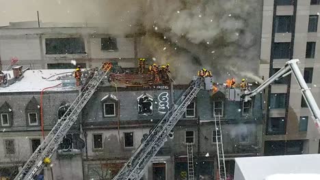 Aerial-shot-looking-down-on-an-apartment-building-as-black-smoke-bellows-from-the-rooftop-as-a-team-of-firefighters-attempt-to-control-the-raging-blaze