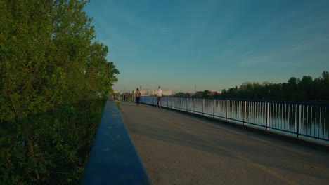 People-walking-on-a-lakeside-pathway-with-railings-and-greenery-at-sunset-in-Jarun-Lake,-Zagreb