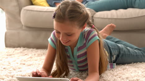 In-Zeitlupe-Glückliches-Mädchen-Mit-Digitalem-Tablet-Auf-Teppich-Mit-Mutter,-Die-Im-Hintergrund-Ein-Buch-Liest