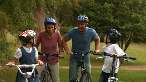 Familia-Feliz-En-Su-Bicicleta-En-El-Parque-Sonriendo-A-La-Cámara