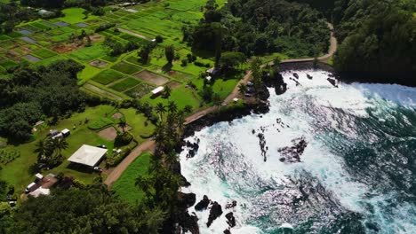 A-drone-shot-of-the-coast-of-Maui,-Hawaii,-as-the-waves-crash-into-the-black-rock-shore,-with-farmland-and-dirt-roads-in-the-background