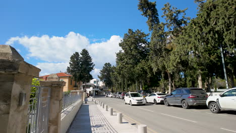 A-street-view-in-Pafos-with-parked-cars,-tall-trees,-and-residential-buildings-under-a-clear-blue-sky