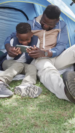 Vertical-video:-Father-and-son,-both-African-American,-are-sitting-by-blue-tent-using-a-tablet
