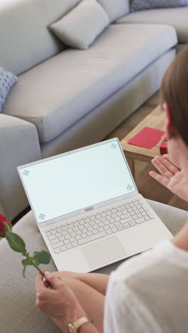 Vertical-video:-Caucasian-woman-holding-red-rose,-using-laptop-at-home