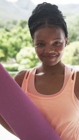 Vídeo-Vertical:-Joven-Afroamericana-Sosteniendo-Una-Estera-De-Yoga,-Sonriendo-Ampliamente-En-Casa