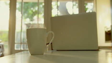 Close-up-of-mug-of-coffee-and-laptop-on-dining-table-in-kitchen-of-comfortable-home-4k