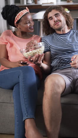 Vertical-video:-Diverse-couple-enjoying-popcorn-at-home,-looking-at-each-other