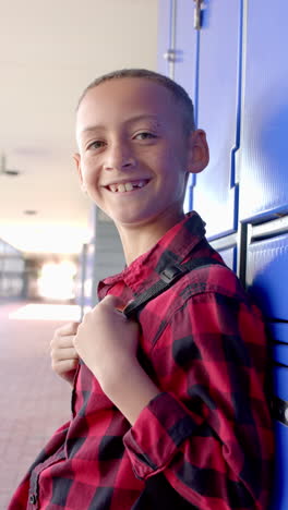 Vertical-video:-In-school,-young-boy-wearing-a-red-checkered-shirt-stands-by-blue-lockers