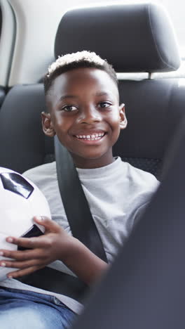 Vídeo-Vertical:-Niño-Afroamericano-Sosteniendo-Una-Pelota-De-Fútbol,-Sonriendo-En-El-Auto