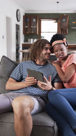 Vertical-video:-Diverse-couple-sitting-on-sofa-at-home,-looking-at-tablet