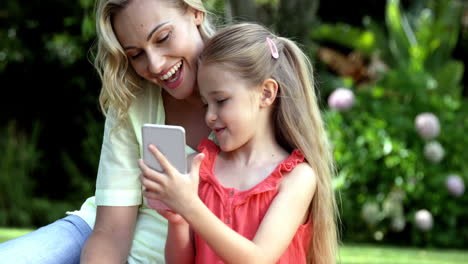 Cute-mother-and-daughter-are-sitting-on-grass-and-looking-a-smartphone