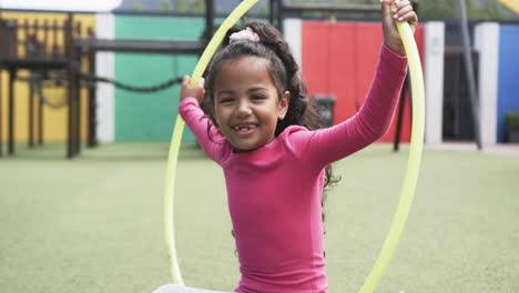 In-a-school-playground,-a-young-African-American-girl-smiles-with-a-hula-hoop