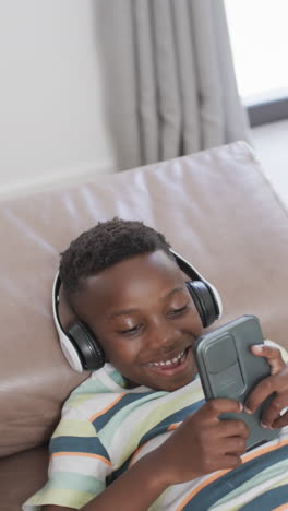Vídeo-Vertical:-Niño-Afroamericano-Usando-Audífonos,-Sosteniendo-Un-Teléfono-Inteligente