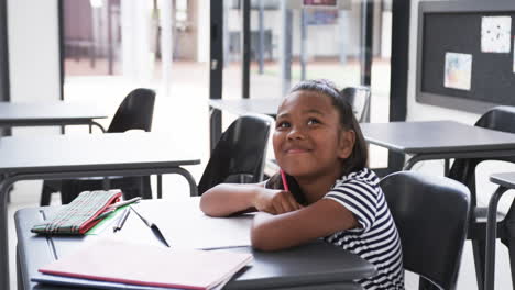 In-a-school-classroom,-a-young-biracial-girl-sits-at-a-desk-looking-up,-with-copy-space