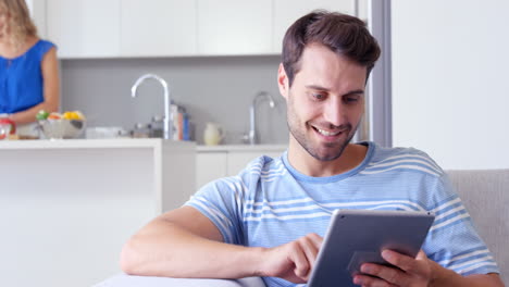 Lächelnder-Mann-Mit-Tablet-Und-Seiner-Frau-Hinter-Ihm-Beim-Zubereiten-Des-Essens