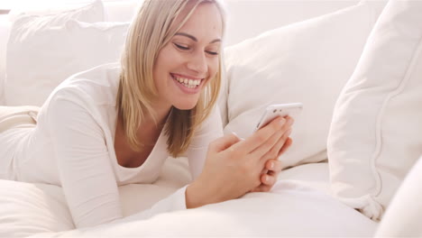 Blonde-Frau-Liegt-Auf-Einem-Sofa,-Benutzt-Ihr-Smartphone-Und-Lächelt