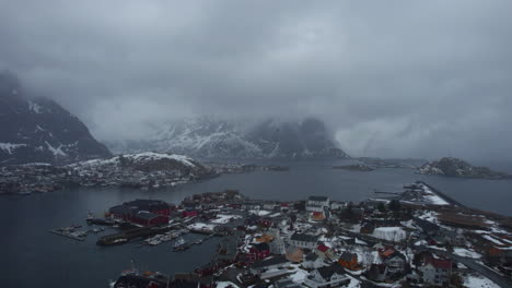Mesmerizing-drone-flight-over-Reine,-Lofoten-Islands,-Norway,-featuring-a-cloudy-sky,-snowy-mountains,-and-a-charming-village-with-homes-and-streets