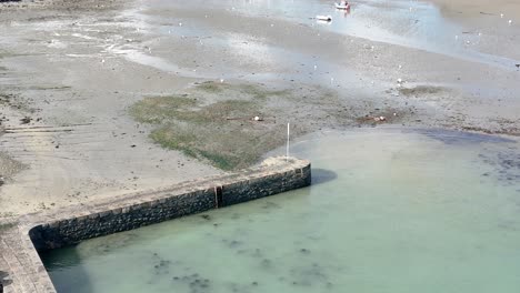 Un-Dron-Que-Cae-Y-Gira-En-Círculos-Sobre-El-Puerto-De-Burdeos,-Guernsey,-Durante-La-Marea-Baja-Con-El-Muro-Del-Puerto,-El-Embarcadero,-Los-Barcos-Secándose-En-La-Arena,-La-Playa-Y-Las-Cabañas-Al-Fondo-En-Un-Día-Brillante