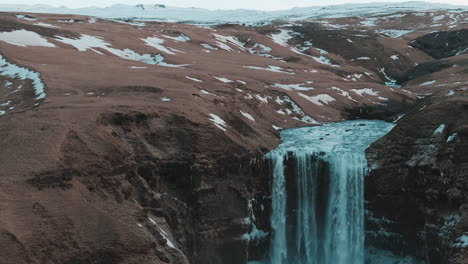 Breathtaking-drone-shot-of-Skógafoss-waterfall-in-Iceland,-showcasing-water-falling-and-a-stunning-snowy-landscape
