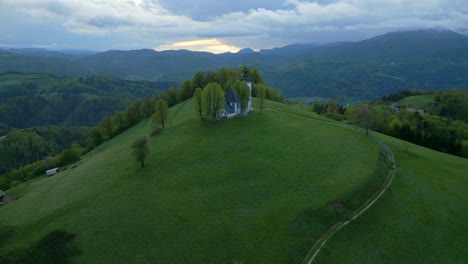 Vista-Aérea-De-Una-Tranquila-Puesta-De-Sol-Sobre-Una-Verde-Colina-Bukov-Vrh-Con-Una-Iglesia-Aislada,-Sensación-De-Paz-Y-Belleza