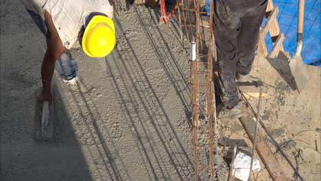 Tilt-down-slow-motion-shot-of-a-mexican-latin-construction-worker-with-a-hardhat-flattening-the-fresh-concrete-using-a-wooden-trowel-on-a-sunny-afternoon