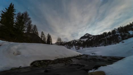 Zeitraffer-Des-Winternachthimmels-Mit-Sternen-über-Bergalpen,-Bach-Und-Schnee-Mit-Dünnen-Wolken