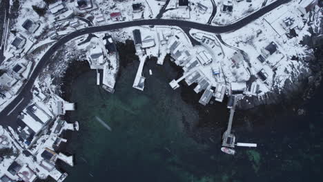 Top-down-drone-shot-of-Reine,-Norway,-covered-in-snow-with-homes,-streets,-and-clear-ocean-water,-as-snow-falls-and-blows-hard-in-the-wind