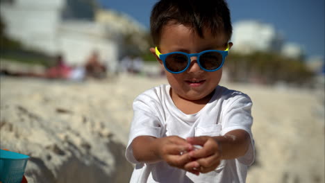 Slow-motion-of-a-cute-young-mexican-latin-boy-wearing-sunglasses-and-a-white-t-shirt-showing-some-seashells-to-the-camera-and-then-putting-them-in-a-bucket