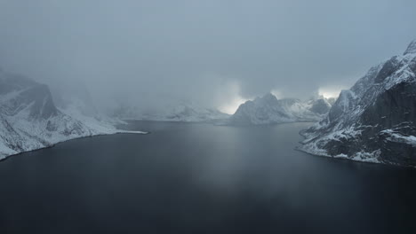 Enchanting-drone-flight-over-the-ocean-in-Reine,-Norway,-featuring-snowfall,-snow-covered-mountains,-and-a-cloudy-sky