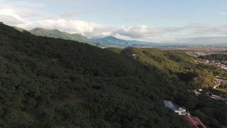 Low-drone-flight-over-tree-tops-on-mountain-side,-mountains-and-cloud-in-distance-sobre-Salta-Capital