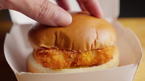 Hand-placing-a-bun-on-a-crispy-fried-fish-sandwich-with-mayonnaise-and-cheese-in-a-fast-food-box