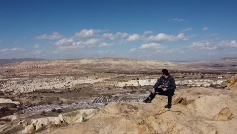 Aventura-En-Capadocia:-Un-Excursionista-Turístico-Solitario-Se-Sienta-En-La-Cima-De-Una-Cueva-De-Montaña-Disfrutando-De-La-Vista-Hacia-Göreme-Y-El-Castillo-De-Uchisar-En-El-Horizonte