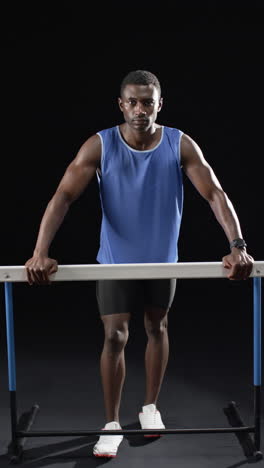 Vertical-video:-African-American-male-athlete-holding-onto-horizontal-hurdle,-black-background