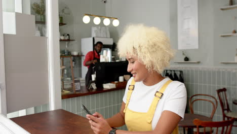 Biracial-woman-with-curly-blonde-hair-uses-a-smartphone-in-a-cafe