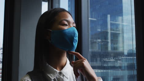 Thoughtful-asian-woman-wearing-face-mask-looking-out-of-window-at-modern-office