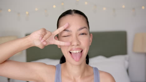 Young-biracial-woman-makes-a-playful-peace-sign-over-her-eye-in-her-bedroom