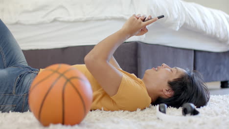 Asian-college-student-lying-on-floor,-holding-smartphone,-basketball-beside-him