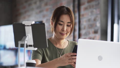 Mujer-Asiática-De-Mediana-Edad-Revisando-El-Teléfono-Inteligente,-Trabajando-Con-Una-Computadora-Portátil