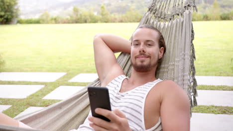 A-young-Caucasian-male-relaxes-in-a-hammock-in-the-backyard-at-home,-smartphone-in-hand