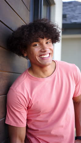 Vertical-video:-A-young-biracial-man-with-a-bright-smile-leans-against-a-wooden-wall