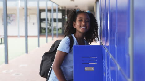 Biracial-girl-with-curly-hair-stands-by-blue-lockers-in-school,-backpack-on-her-shoulders