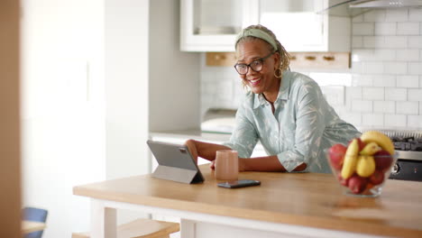 African-American-senior-woman-using-tablet-in-kitchen,-smiling-at-screen