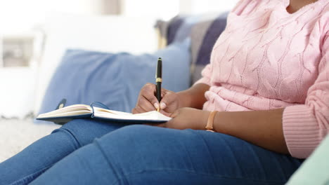 Eine-Afroamerikanische-Frau-Schreibt-In-Ein-Notizbuch,-Während-Sie-Zu-Hause-Auf-Einer-Couch-Sitzt