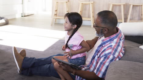 Biracial-grandfather-and-granddaughter-sitting,-holding-tablet